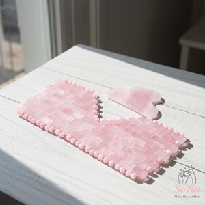 May include: A pink rose quartz eye mask and a heart-shaped gua sha tool. The eye mask is made of small, rectangular rose quartz pieces and pink beads. The gua sha tool is smooth and polished. The items are on a white wooden surface.