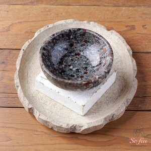 Puede incluir: Un peque&ntilde;o cuenco de piedra moteada con interior oscuro se asienta sobre un bloque blanco, colocado dentro de un plato de piedra m&aacute;s grande y de color claro. El cuenco tiene una superficie pulida con motas rojas, negras y blancas. El plato tiene un borde rugoso y texturizado.