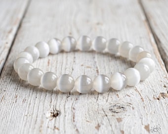 May include: A stretch bracelet composed of smooth, round, light gray beads with a chatoyant effect. Each bead displays a subtle, luminous band of light running through its center, creating a soft, reflective appearance across the entire circular piece.