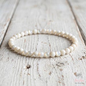 May include: A circular bracelet composed of small, smooth, off-white beads with a subtle pearlescent sheen. The beads are uniform in size and arranged in a continuous loop, reflecting light across their polished, rounded surfaces.