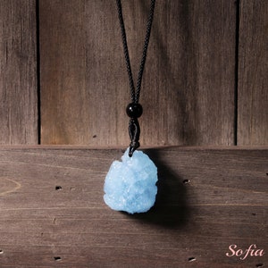 Puede incluir: Un colgante de cristal azul claro en un collar de cord&oacute;n negro. El colgante tiene una forma irregular y rugosa y tiene un aspecto brillante y transl&uacute;cido.