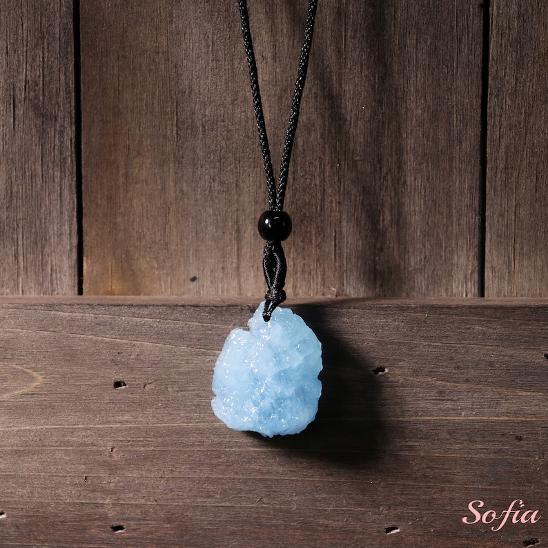 Puede incluir: Un colgante de cristal azul claro en un collar de cord&oacute;n negro. El colgante tiene una forma irregular y rugosa y tiene un aspecto brillante y transl&uacute;cido.