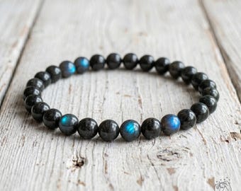 May include: A circular beaded bracelet composed of smooth, dark grey labradorite stones. Each spherical bead features a polished finish, with select stones displaying iridescent blue flashes of light across their dark, reflective surfaces.