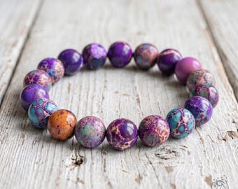 Pode incluir: Uma pulseira elástica composta por contas redondas e polidas em vários tons de roxo, azul-petróleo e laranja. As contas apresentam uma textura de pedra natural rachada com padrões de veios intrincados, dispostas em círculo.