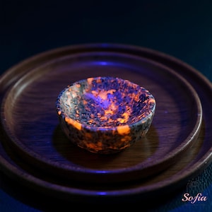 Puede incluir: Un peque&ntilde;o cuenco redondo con una superficie moteada y texturizada, que muestra vibrantes tonos naranja y azul. El cuenco se encuentra sobre un plato de madera, creando un contraste de texturas y colores. El interior del cuenco brilla con luz propia.