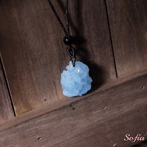 May include: A light blue crystal pendant on a black cord necklace. The crystal is a cluster of small, irregular shapes.