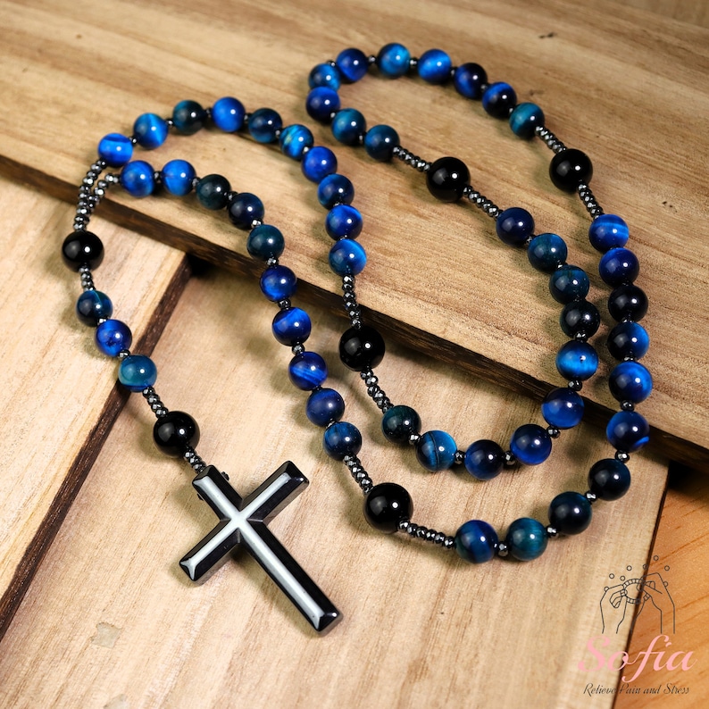 Pu&ograve; includere: Un rosario con collana di perline blu e nere con un ciondolo a croce in metallo. Le perline sono in occhio di tigre.