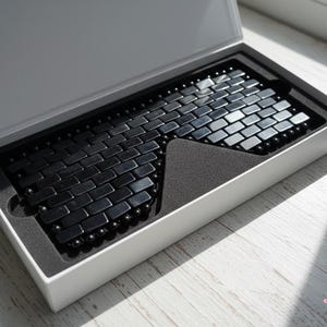 May include: A black magnetic bracelet, made of rectangular and spherical beads, is displayed in a white box with a black interior. The bracelet's design resembles a brick pattern. The box is open, revealing the bracelet.