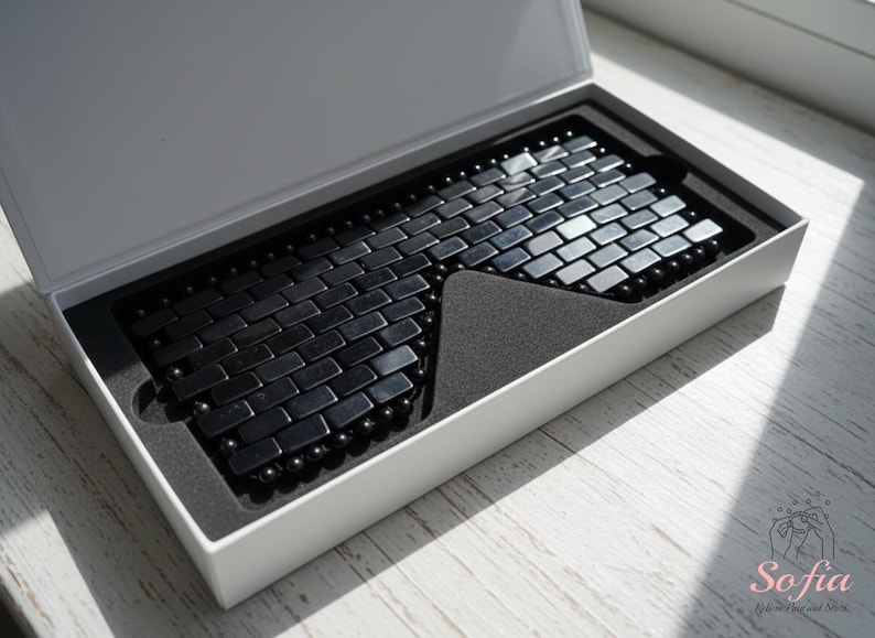 May include: A black magnetic bracelet, made of rectangular and spherical beads, is displayed in a white box with a black interior. The bracelet's design resembles a brick pattern. The box is open, revealing the bracelet.