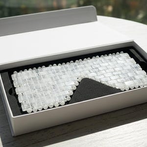 May include: A clear, rectangular stone eye mask in an open white box with a black interior. The mask is designed to cover the eyes, possibly for relaxation or beauty treatments. The box is on a white table.