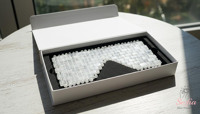 May include: A clear, rectangular stone eye mask in an open white box with a black interior. The mask is designed to cover the eyes, possibly for relaxation or beauty treatments. The box is on a white table.