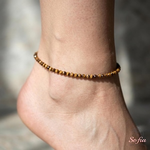 May include: An anklet composed of small, round, faceted tiger's eye beads in shades of golden brown and dark brown. The beads are strung together in a single continuous line, creating a delicate and uniform circular band around the ankle.
