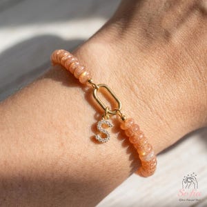 Peut inclure: Un bracelet composé de perles rondes en pierre de soleil couleur pêche avec un fermoir ovale doré. Une petite breloque en forme de lettre S dorée, incrustée de minuscules pierres transparentes, est suspendue au centre du fermoir.