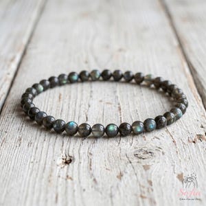 Puede incluir: Una pulsera de cuentas compuesta por pequeñas piedras redondas de labradorita gris oscuro. Cada piedra presenta un acabado liso y pulido con sutiles destellos iridiscentes azules y verdes visibles en la superficie de las cuentas dispuestas en círculo.