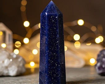 May include: A tall, polished blue sandstone obelisk with sparkling inclusions. The gemstone is a deep, rich blue with a pointed top. The obelisk is standing on a gray surface. Decorative item.