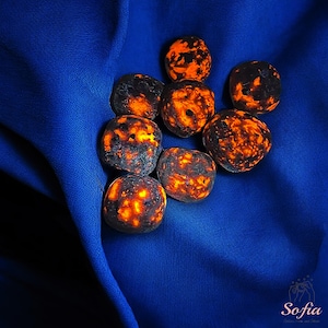 May include: A group of eight smooth, round, dark gray stones with orange and red inclusions. The stones are arranged on a blue fabric background.