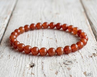 May include: A circular stretch bracelet composed of smooth, polished, round carnelian beads in varying shades of translucent orange and deep reddish-brown. The beads are uniform in size and strung together to form a continuous loop.