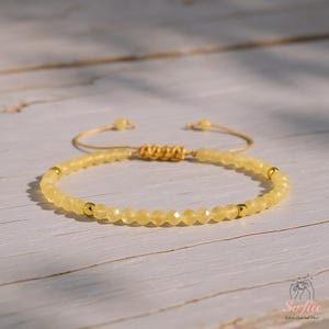 May include: A yellow beaded bracelet with gold accents. The bracelet features small, faceted beads and gold-colored metal spacers. The adjustable cord allows for a custom fit. The bracelet is displayed on a light-colored wooden surface.