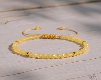 May include: A yellow beaded bracelet with gold accents. The bracelet features small, faceted beads and gold-colored metal spacers. The adjustable cord allows for a custom fit. The bracelet is displayed on a light-colored wooden surface.