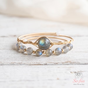 May include: A gold-toned wire-wrapped bracelet featuring a central round labradorite gemstone and a second strand composed of smaller round labradorite beads interspersed with tiny gold-toned metal spheres. The logo Sofia Relieve Pain and Stress is visible.