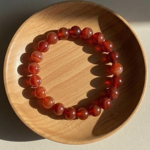 May include: A circular bracelet composed of polished, translucent red-orange agate beads. Each spherical bead features subtle, concentric banding patterns in varying shades of deep amber and rust, arranged in a continuous loop on an elastic cord.