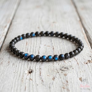 May include: A beaded bracelet composed of small, round, dark grey labradorite stones. The beads feature a smooth, polished finish with iridescent blue flashes visible on several stones. The bracelet is arranged in a circular shape.