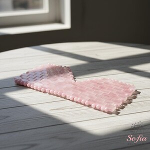 May include: A pink rose quartz eye mask. The mask is constructed from small, rectangular stones and is shaped to fit around the eyes. It rests on a light-coloured wooden surface.