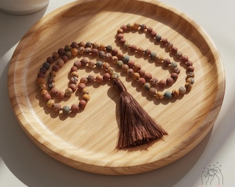 May include: A beaded necklace with a brown tassel rests on a wooden tray. The necklace features small, round beads in shades of brown, tan, and a touch of green. The tassel is a darker shade of brown. A white ceramic cup is in the background.