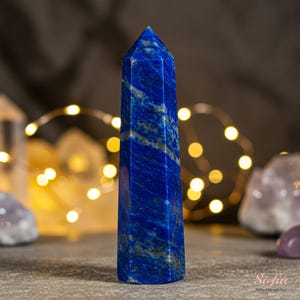 May include: A deep blue lapis lazuli crystal point, also known as a wand, with a pointed tip and a smooth, polished surface. The crystal is set against a background of warm, golden lights.