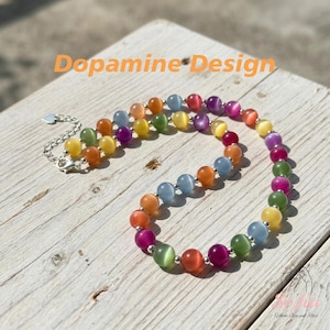 May include: A beaded necklace featuring round, multi-colored cat's eye beads in shades of orange, yellow, green, blue, purple, and pink, separated by small silver-toned metal spacers, with a silver-toned chain extender and the text Dopamine Design.