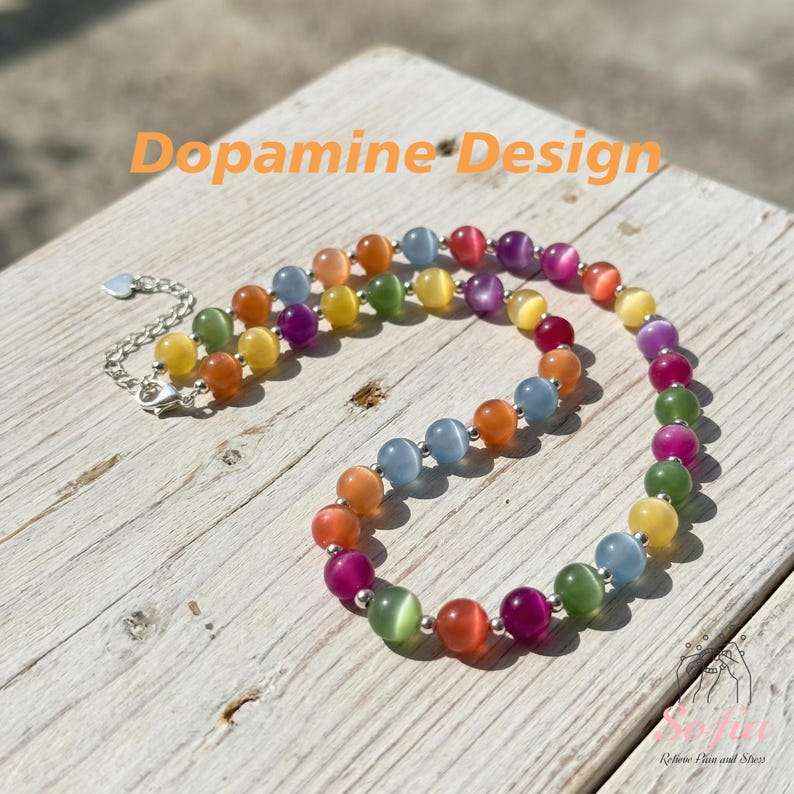 May include: A beaded necklace featuring round, multi-colored cat's eye beads in shades of orange, yellow, green, blue, purple, and pink, separated by small silver-toned metal spacers, with a silver-toned chain extender and the text Dopamine Design.
