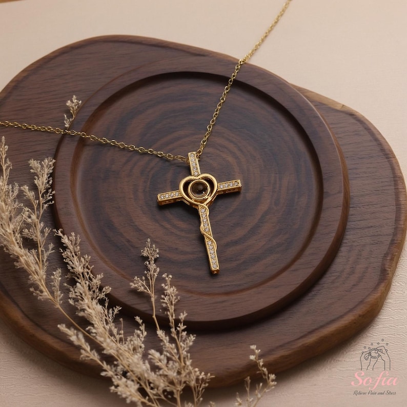 Puede incluir: Un collar dorado con un colgante de cruz, que incorpora un dise&ntilde;o en forma de coraz&oacute;n en el centro. La cruz est&aacute; adornada con peque&ntilde;as piedras brillantes. El collar descansa sobre una bandeja de madera oscura, con una ramita de flores secas en primer plano. La cadena es fina.