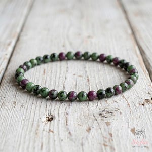 May include: A beaded bracelet featuring small, round, polished stones in alternating shades of deep magenta, forest green, and black. The beads are strung together in a continuous circular loop, showcasing natural color variations and textures.