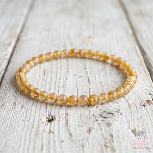 May include: A circular bracelet composed of small, polished, translucent golden-yellow beads. The beads are uniform in size and shape, featuring internal inclusions and a reflective, glossy finish that catches the light, arranged in a continuous loop.
