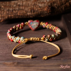 May include: A gold beaded bracelet with a heart-shaped charm made of colourful jasper. The bracelet has a sliding knot closure.