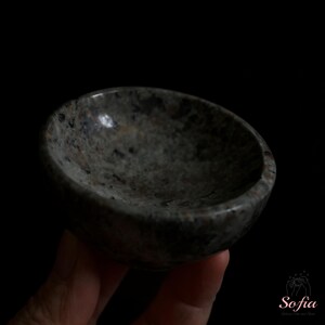 Puede incluir: Un peque&ntilde;o cuenco redondo de piedra con un patr&oacute;n moteado gris y negro. El cuenco se sostiene en una mano sobre un fondo negro. El interior del cuenco es liso y el exterior tiene una textura rugosa.