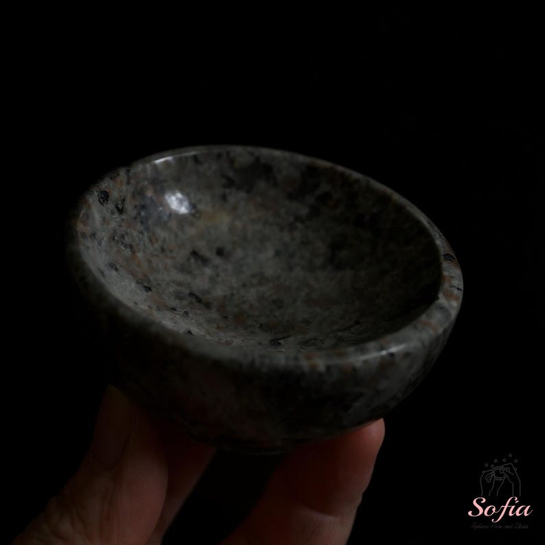Puede incluir: Un peque&ntilde;o cuenco redondo de piedra con un patr&oacute;n moteado gris y negro. El cuenco se sostiene en una mano sobre un fondo negro. El interior del cuenco es liso y el exterior tiene una textura rugosa.