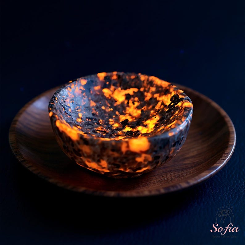 Puede incluir: Un cuenco peque&ntilde;o, redondo y moteado, con tonos naranja y negro, se encuentra sobre un plato de madera. El cuenco tiene una superficie rugosa y texturizada y parece brillar con una luz interior. El plato de madera es de color marr&oacute;n c&aacute;lido.