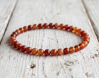 May include: A circular beaded bracelet composed of small, polished, spherical carnelian stones in varying shades of translucent orange, deep red, and burnt amber, arranged in a continuous loop on a thin, flexible, and elastic cord.