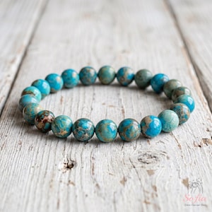 May include: A stretch bracelet composed of round, polished beads in varying shades of turquoise, teal, and brown. The beads feature natural veining and marbled patterns throughout, arranged in a circular loop on a light-colored wooden surface.