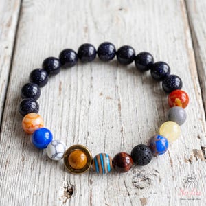 Peut inclure: Un bracelet élastique composé de perles rondes représentant le système solaire. Le design comprend des perles bleu foncé pailletées, une perle blanche marbrée, une perle jaune, une perle grise, une perle rouge et une perle centrale avec un anneau doré.
