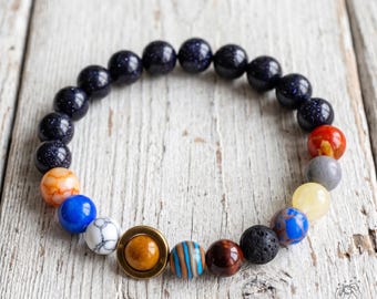 Könnte beinhalten: Ein elastisches Armband mit runden Perlen, die das Sonnensystem darstellen. Das Design umfasst dunkelblaue Glitzerperlen, eine weiß marmorierte Perle, eine gelbe Perle, eine graue Perle, eine rote Perle und eine mittlere Perle mit einem goldenen Ring.