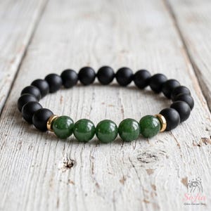 May include: A beaded bracelet featuring matte black spherical beads and five polished green jade-like beads in the center. The green beads are flanked by small gold-toned metal spacers. The bracelet is arranged in a circular shape on a light surface.