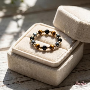 May include: A beaded ring featuring alternating round tiger eye, black onyx, and faceted hematite beads separated by small gold-toned metal spacers. The ring is arranged in a circular band pattern with a mix of brown, black, and metallic gray tones.