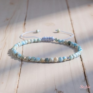 May include: A light blue beaded bracelet with a sliding knot fastening. The bracelet features small, round beads in shades of blue, white, and grey. The bracelet is displayed on a light-coloured wooden surface.