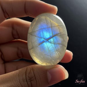May include: A smooth, oval-shaped moonstone with a blue and iridescent sheen. The stone is held in a hand.