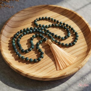 May include: A dark green beaded necklace with a beige tassel, displayed on a round wooden tray. The beads are a deep, mottled green, and the tassel is a light beige colour. The necklace is arranged in a circular pattern.