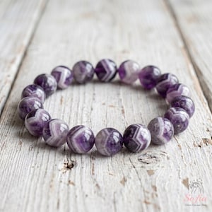 Peut inclure: Un bracelet élastique composé de perles d'améthyste rondes et polies. Chaque perle présente un motif en chevron distinct avec des bandes alternées de violet profond et de blanc translucide, créant un effet rayé uniforme sur tout le bijou.