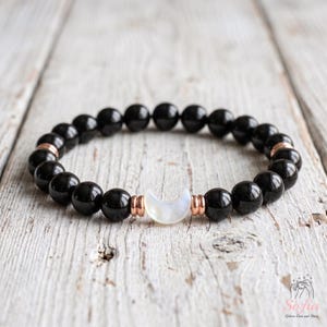 May include: A beaded bracelet featuring smooth, round black beads arranged in a circular pattern. The bracelet includes a central white crescent moon charm flanked by two small rose gold-toned metal spacers on each side of the moon.