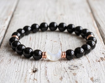 May include: A beaded bracelet featuring smooth, round black beads arranged in a circular pattern. The bracelet includes a central white crescent moon charm flanked by two small rose gold-toned metal spacers on each side of the moon.